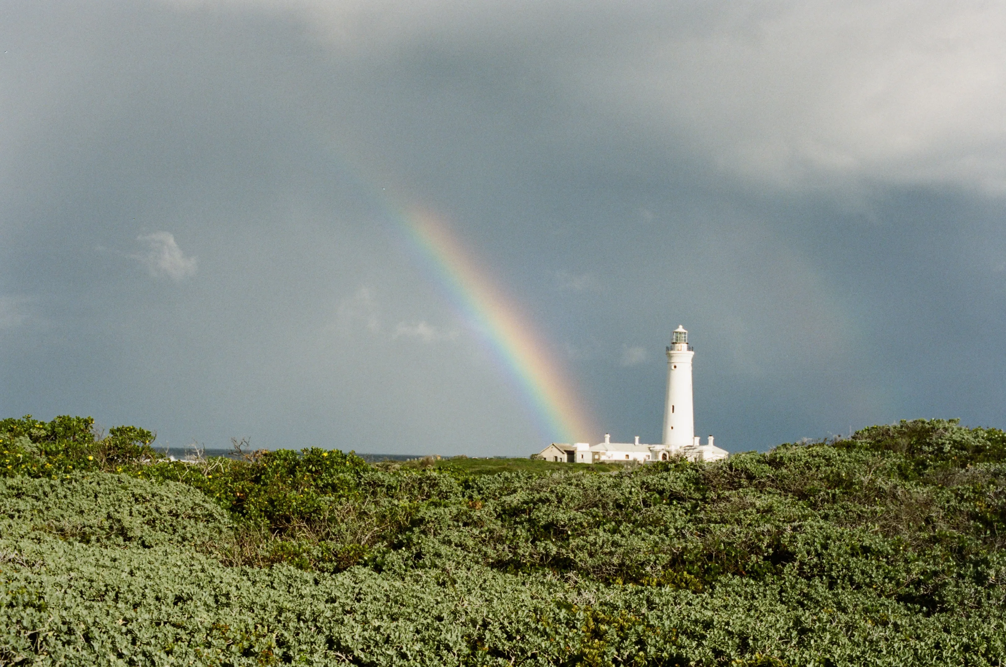 Seal Point Lighthouse under a storm sky, a rainbow falling towards it over low Fynbos.